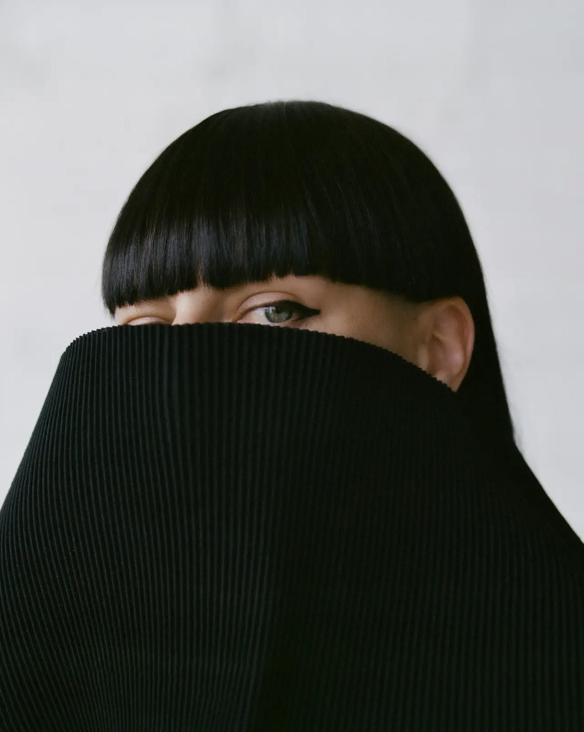 A close-up portrait of MELITTA BAUMEISTER with straight black hair and bangs, partially covered by a black pleated fabric face veil, against a light gray background.