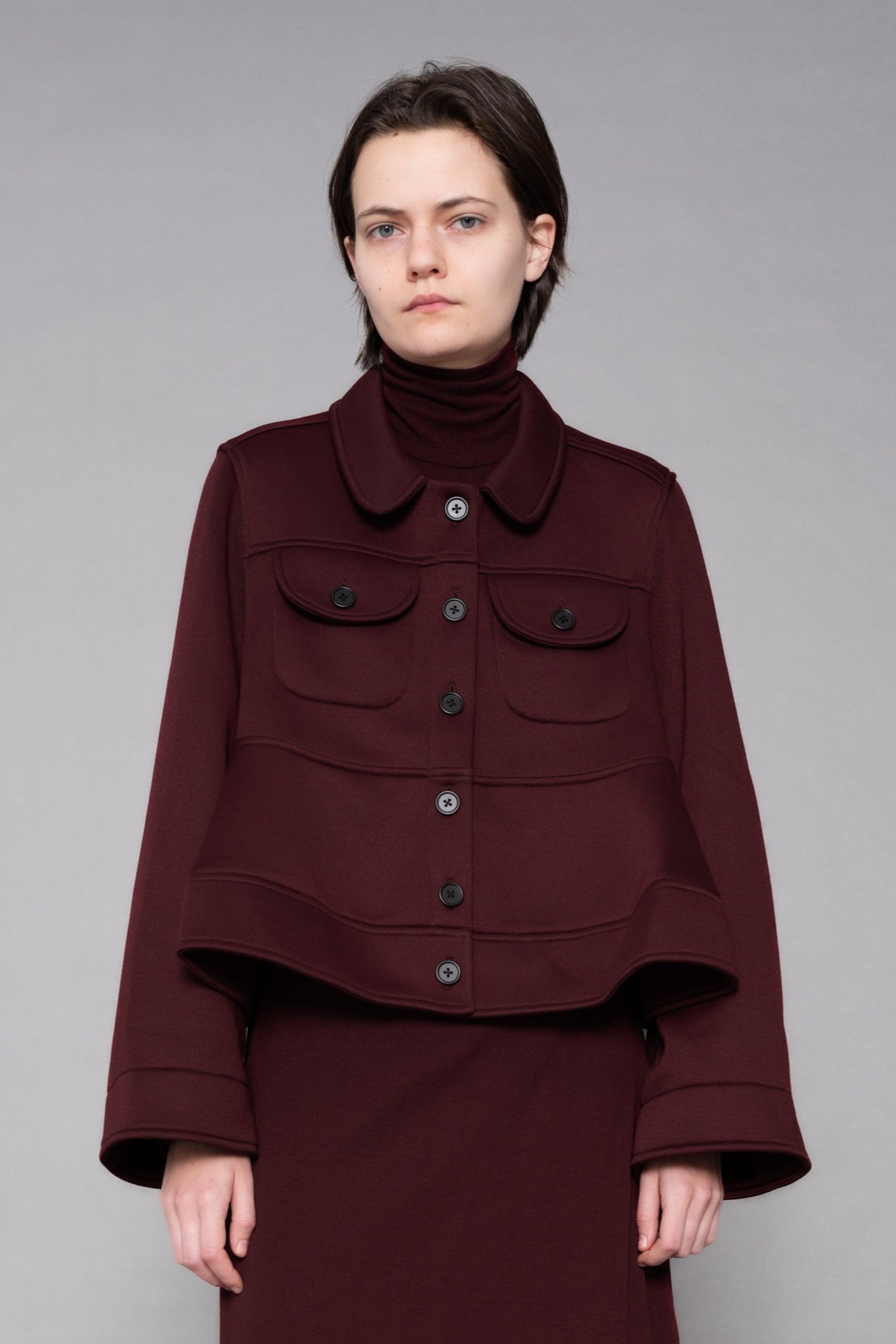 Person wearing a burgundy outfit with a jacket and skirt on a gray background