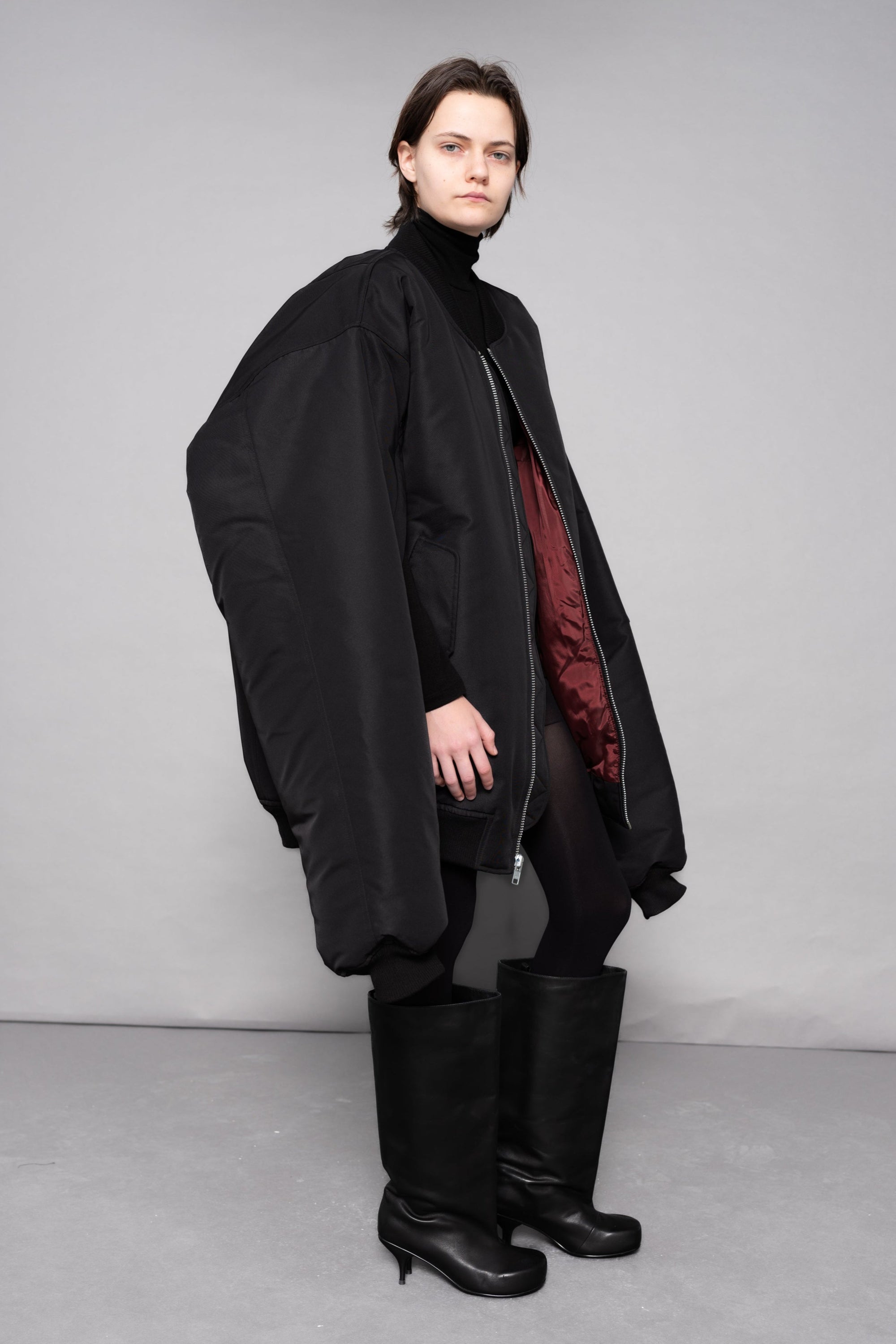 Person wearing a black coat and boots against a gray background