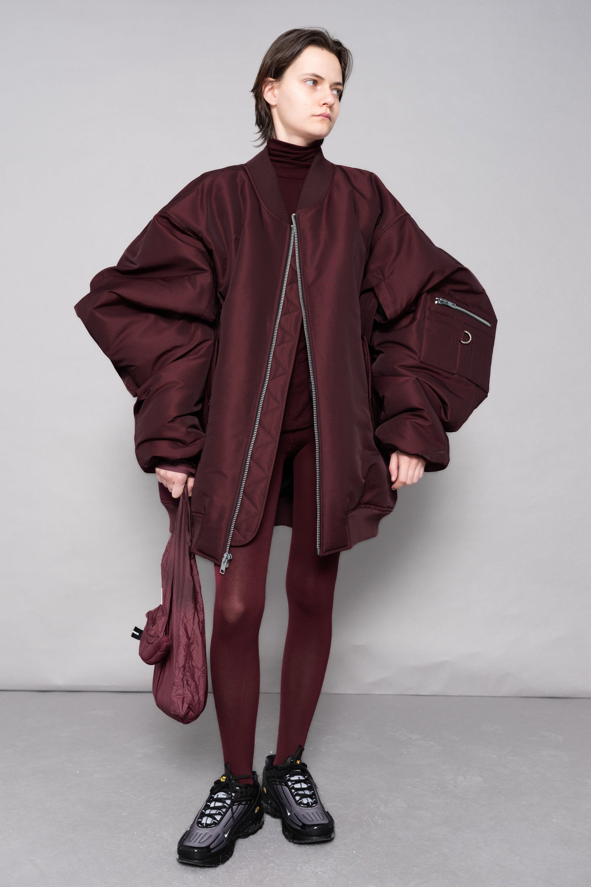 Person wearing a burgundy outfit with a matching jacket and pants, holding a matching bag against a gray background.