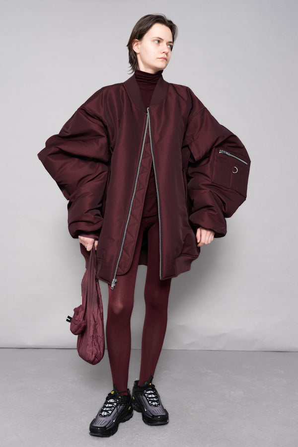 Person wearing a burgundy outfit with a matching jacket and pants, holding a matching bag against a gray background.