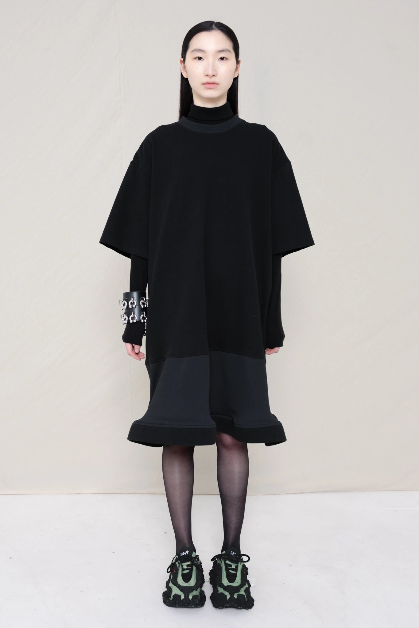 woman-wearing-A-black-T-SHIRT-STYLE-DRESS-WITH-A-WAVY-HEM-over-a-black-turtle-neck-paired-with-custom-shoes-and-black-leather-studded-bracelet-against-a-plain-background-full-body-front-view