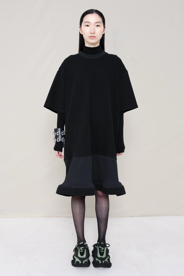 woman-wearing-A-black-T-SHIRT-STYLE-DRESS-WITH-A-WAVY-HEM-over-a-black-turtle-neck-paired-with-custom-shoes-and-black-leather-studded-bracelet-against-a-plain-background-full-body-front-view