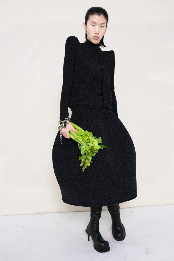 PERSON-WEARING-GLITTER-BLACK-FOAMY-SHOULDER-CARDIGAN-WITH-MATCHING-MOCK-NECK-SHIRT-AND-FOAMY-BALL-SKIRT-WEARING-BLACK-DOUBLE-HEELED-BOOTS-ACCESSORIZED-ON-THE-WRIST-MB-CHARM-BRACELET-AND-HOLDING-CELERY-STALKS-FRONT-VIEW