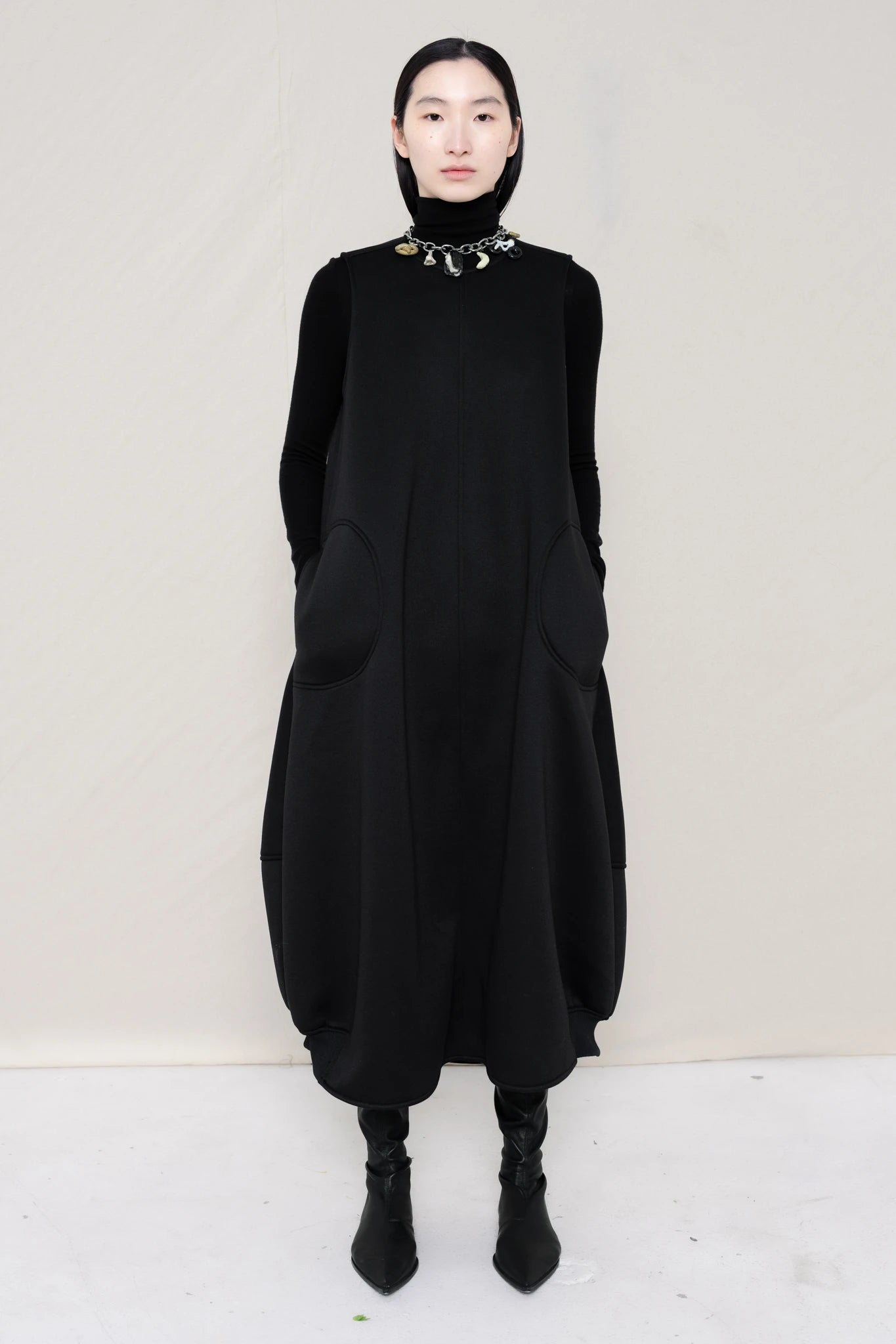 woman-wearing-a-black-SLEEVELESS-O-LINE-SHAPED-DRESS-WITH-round-POCKETS-over-a-black-turtle-neck-black-leather-pointed-boots-and-charm-chain-chocker-against a-white-background-full-body-front-view