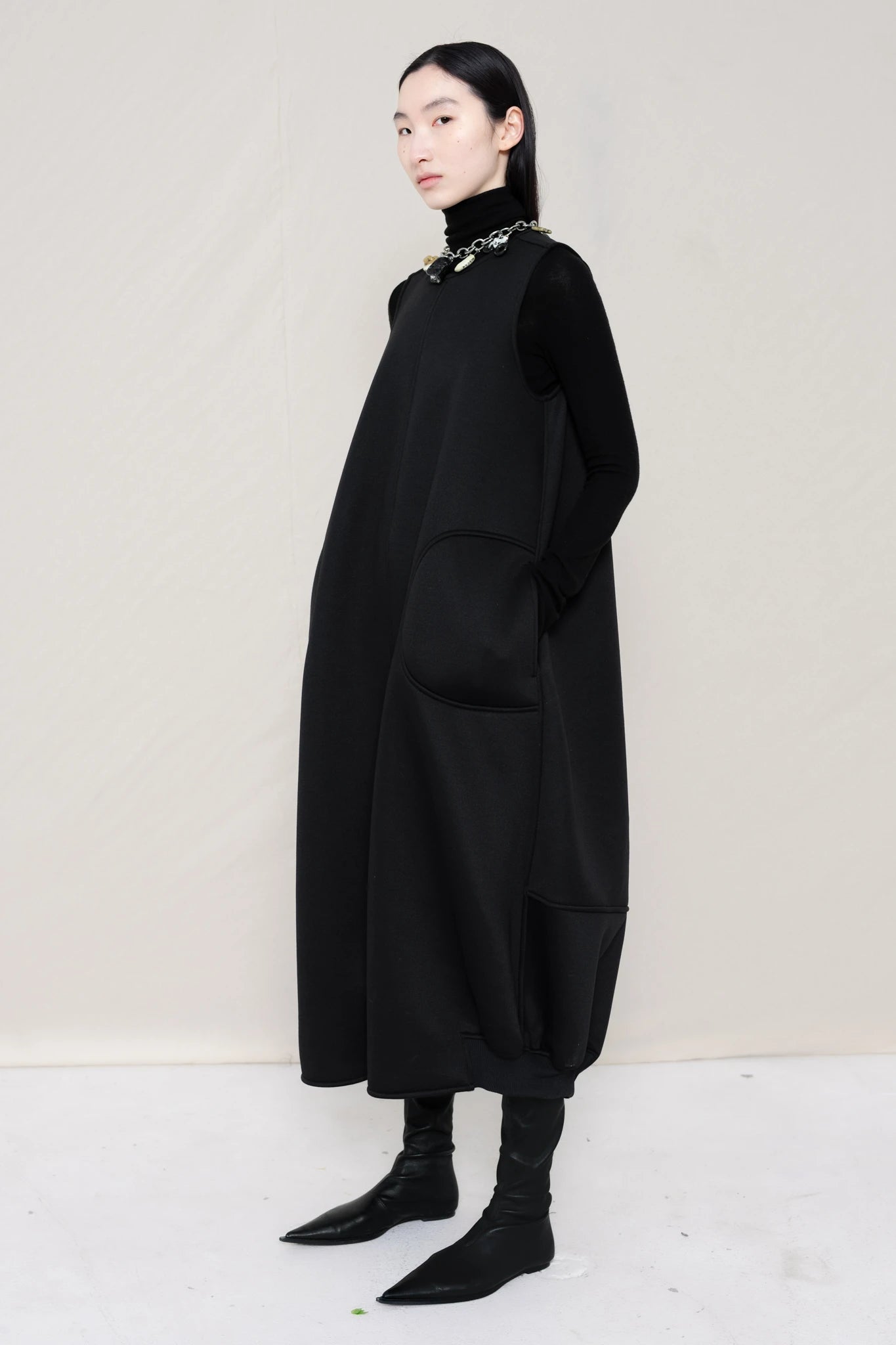 woman-wearing-a-black-SLEEVELESS-O-LINE-SHAPED-DRESS-WITH-round-POCKETS-over-a-black-turtle-neck-black-leather-pointed-boots-and-charm-chain-chocker-against a-white-background-full-body-three-quarters-view