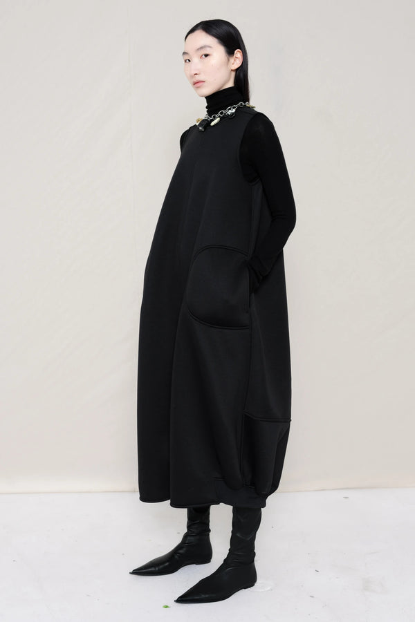 woman-wearing-a-black-SLEEVELESS-O-LINE-SHAPED-DRESS-WITH-round-POCKETS-over-a-black-turtle-neck-black-leather-pointed-boots-and-charm-chain-chocker-against a-white-background-full-body-three-quarters-view