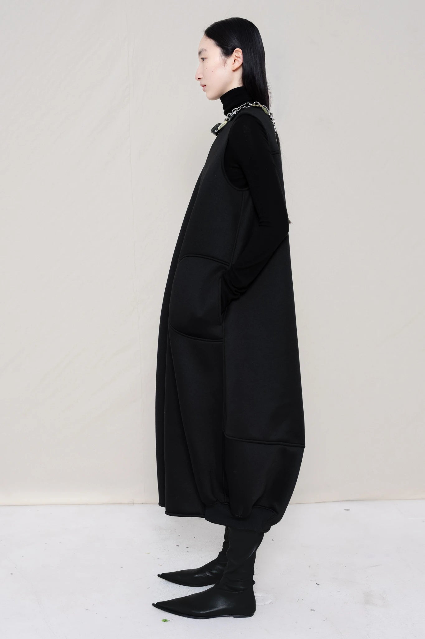 woman-wearing-a-black-SLEEVELESS-O-LINE-SHAPED-DRESS-WITH-round-POCKETS-over-a-black-turtle-neck-black-leather-pointed-boots-and-charm-chain-chocker-against a-white-background-full-body-side-view
