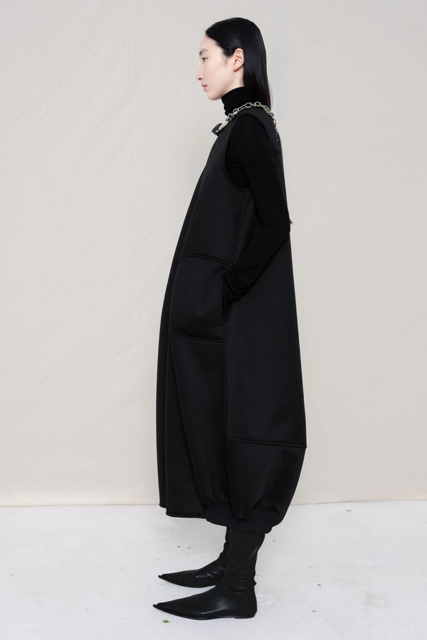 woman-wearing-a-black-SLEEVELESS-O-LINE-SHAPED-DRESS-WITH-round-POCKETS-over-a-black-turtle-neck-black-leather-pointed-boots-and-charm-chain-chocker-against a-white-background-full-body-side-view