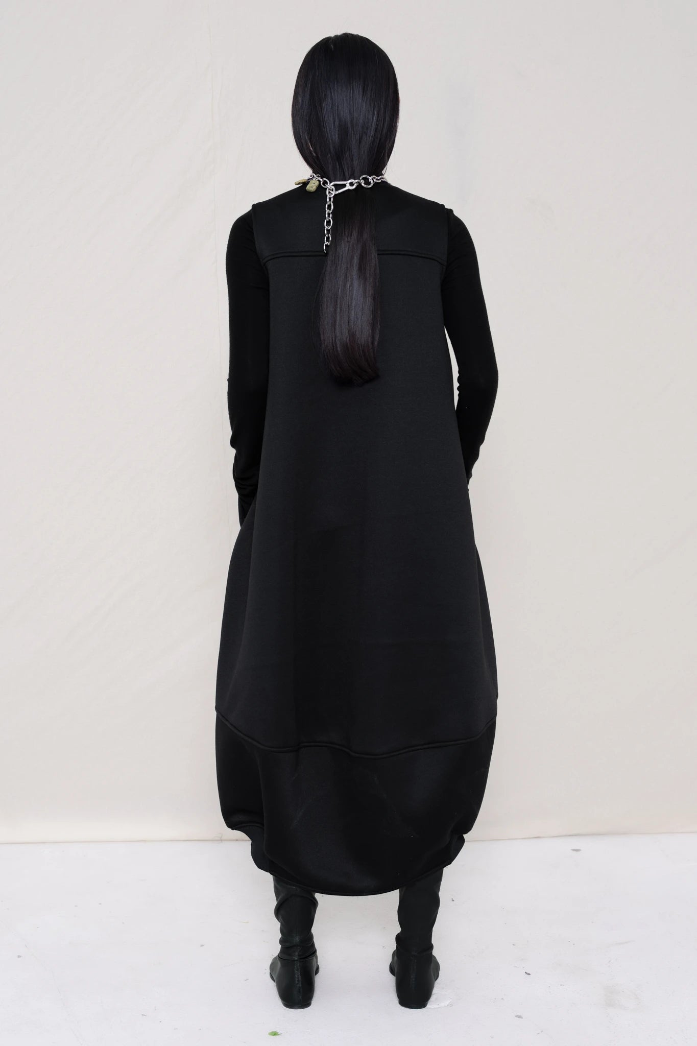 woman-wearing-a-black-SLEEVELESS-O-LINE-SHAPED-DRESS-WITH-round-POCKETS-over-a-black-turtle-neck-black-leather-pointed-boots-and-charm-chain-chocker-against a-white-background-full-body-back-view