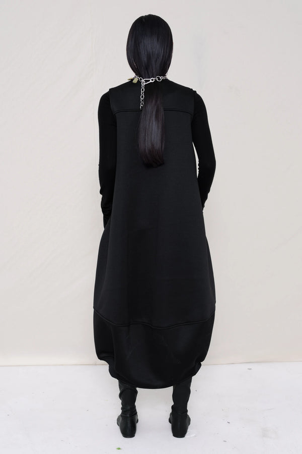 woman-wearing-a-black-SLEEVELESS-O-LINE-SHAPED-DRESS-WITH-round-POCKETS-over-a-black-turtle-neck-black-leather-pointed-boots-and-charm-chain-chocker-against a-white-background-full-body-back-view