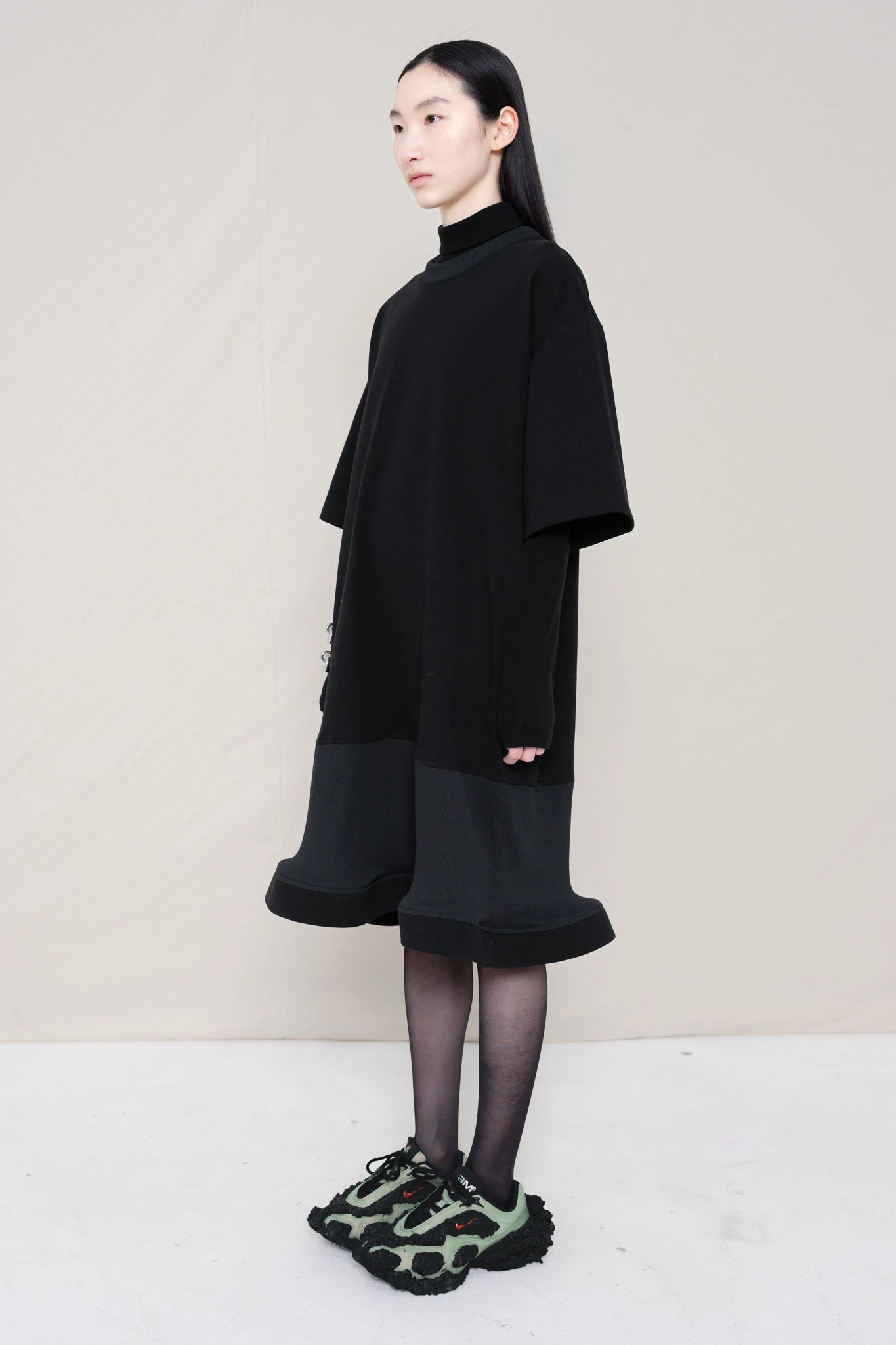 woman-wearing-A-black-T-SHIRT-STYLE-DRESS-WITH-A-WAVY-HEM-over-a-black-turtle-neck-paired-with-custom-shoes-and-black-leather-studded-bracelet-against-a-plain-background-full-body-three-quarters-view