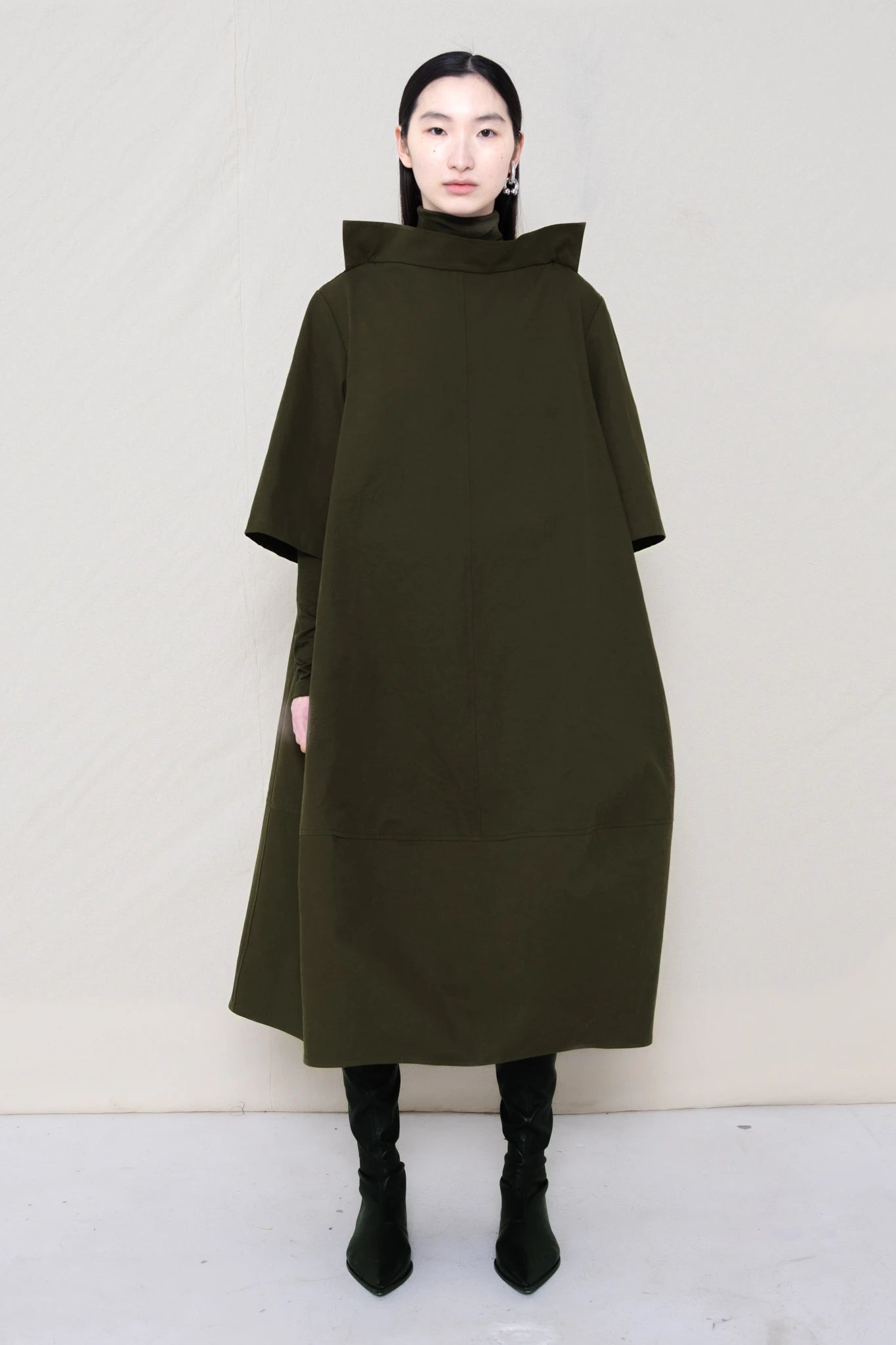 woman-wearing-a-calf-length-moss-green-a-line-boatneck-dress-with-mid-length-sleeves-worn-over-a-moss-green-turtle-neck-worn-with-black-pointed-leather-boots-against-a-plain-background-full-body-front-view