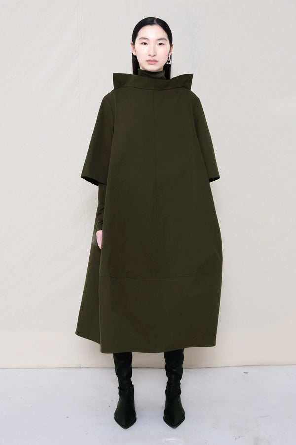 woman-wearing-a-calf-length-moss-green-a-line-boatneck-dress-with-mid-length-sleeves-worn-over-a-moss-green-turtle-neck-worn-with-black-pointed-leather-boots-against-a-plain-background-full-body-front-view
