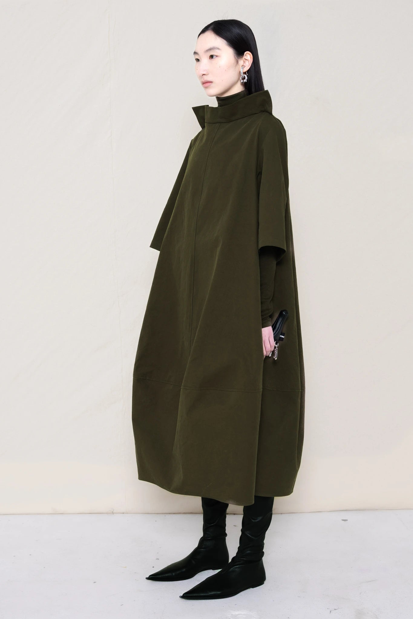 woman-holding-a-black-leather-studded-banana-wearing-a-calf-length-moss-green-a-line-boatneck-dress-with-mid-length-sleeves-worn-over-a-moss-green-turtle-neck-worn-with-black-pointed-leather-boots-against-a-plain-background-full-body-three-quarters-view