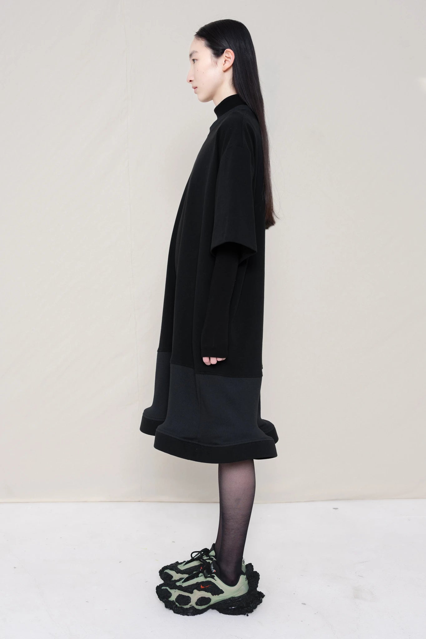 woman-wearing-A-black-T-SHIRT-STYLE-DRESS-WITH-A-WAVY-HEM-over-a-black-turtle-neck-paired-with-custom-shoes-and-black-leather-studded-bracelet-against-a-plain-background-full-body-side-view