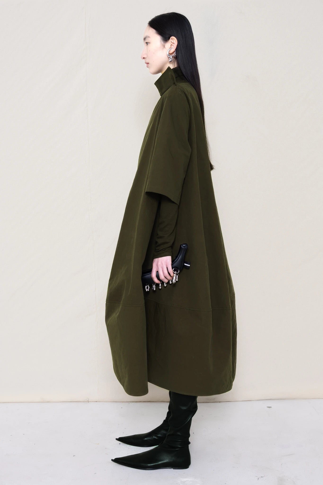 woman-holding-a-black-leather-studded-banana-wearing-a-calf-length-moss- green-a-line-boatneck-dress-with-mid-length-sleeves-worn-over-a-moss-green-turtle-neck-worn-with-black-pointed-leather-boots-against-a-plain-background-full-body-side-view