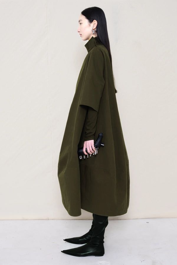 woman-holding-a-black-leather-studded-banana-wearing-a-calf-length-moss- green-a-line-boatneck-dress-with-mid-length-sleeves-worn-over-a-moss-green-turtle-neck-worn-with-black-pointed-leather-boots-against-a-plain-background-full-body-side-view