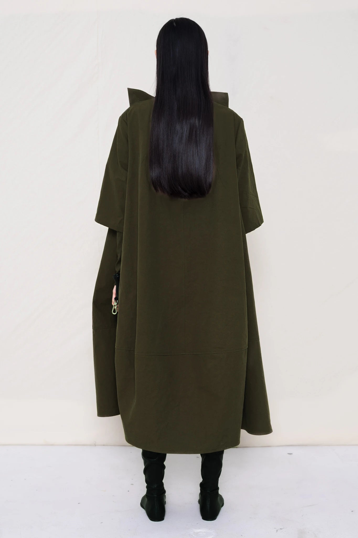 woman-holding-a-black-leather-studded-banana-wearing-a-calf-length-moss-green-a-line-boatneck-dress-with-mid-length-sleeves-worn-over-a-moss-green-turtle-neck-worn-with-black-pointed-leather-boots-against-a-plain-background-full-body-back-view