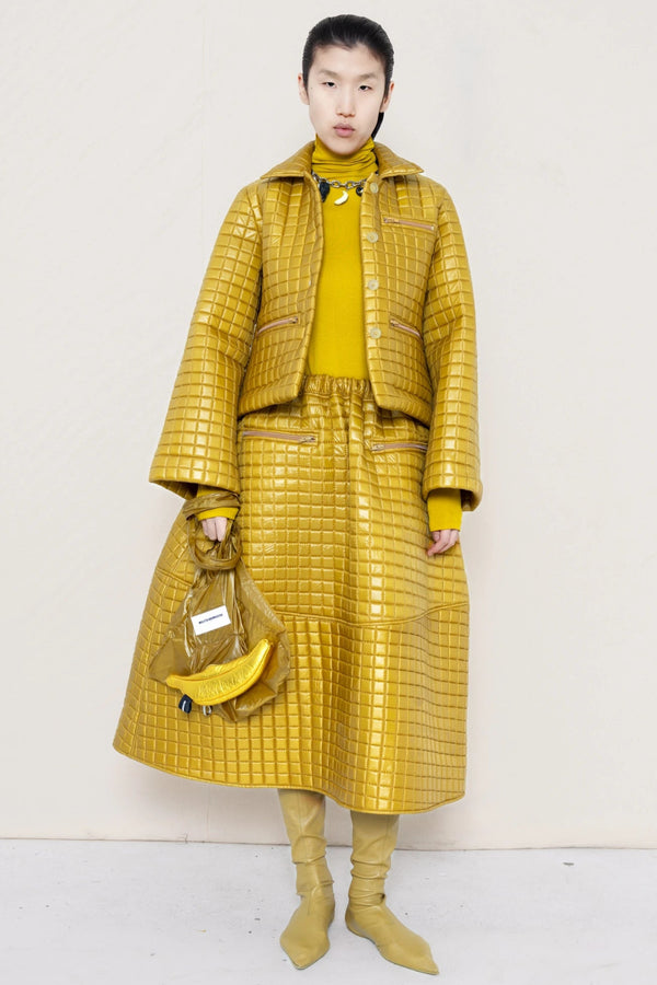 PERSON-WEARING-GOLD-YELLOW-SLEEVE-MOCK-NECK-TOP-WITH-MATCHING-QUILTEDJACKET-WITH-COLLAR-AND-A-LINE-SKIRT-ACCESSORIZED-WITH-MB-CHARM-NECKLACE-AND-MUD-COLORED-POINTY-FLAT-BOOTS-HOLDING--MATCHING-BANANA-BAG-FRONT-VIEW