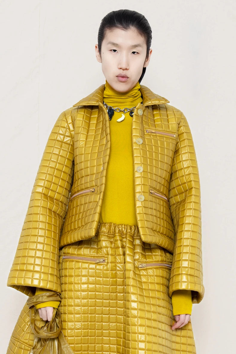 PERSON-WEARING-GOLD-YELLOW-SLEEVE-MOCK-NECK-TOP-WITH-MATCHING-QUILTEDJACKET-WITH-COLLAR-AND-A-LINE-SKIRT-ACCESSORIZED-WITH-MB-CHARM-NECKLACE-AND-MUD-COLORED-POINTY-FLAT-BOOTS-HOLDING--MATCHING-BANANA-BAG-FRONT-VIEW