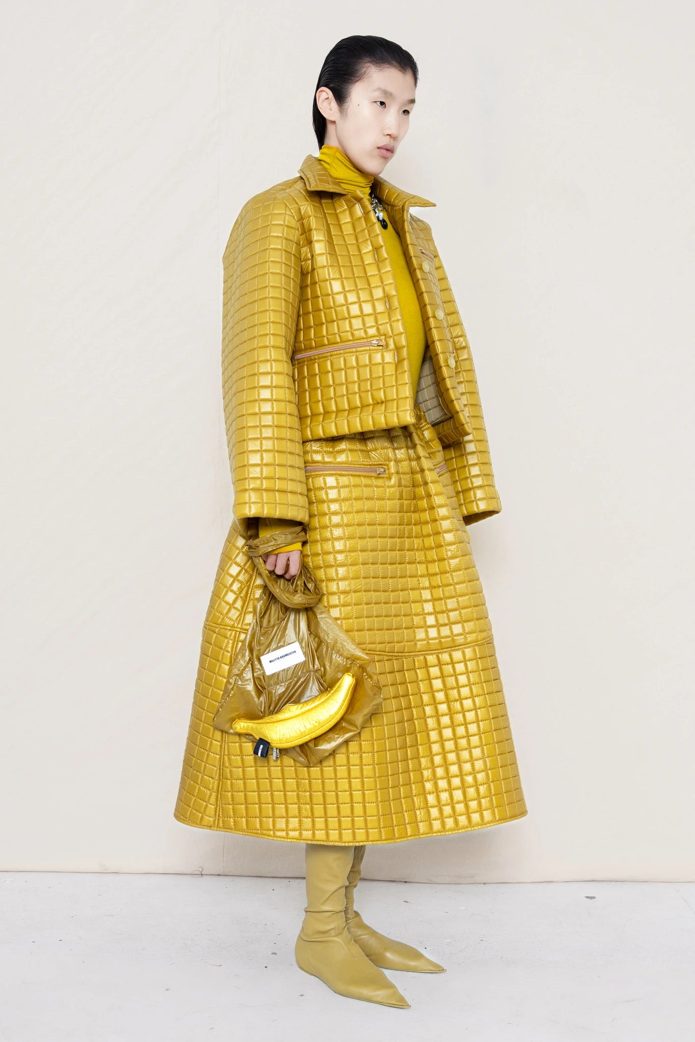 PERSON-WEARING-GOLD-YELLOW-SLEEVE-MOCK-NECK-TOP-WITH-MATCHING-QUILTEDJACKET-WITH-COLLAR-AND-A-LINE-SKIRT-ACCESSORIZED-WITH-MB-CHARM-NECKLACE-AND-MUD-COLORED-POINTY-FLAT-BOOTS-HOLDING--MATCHING-BANANA-BAG-DETAIL-QUATER-FRONT-VIEW