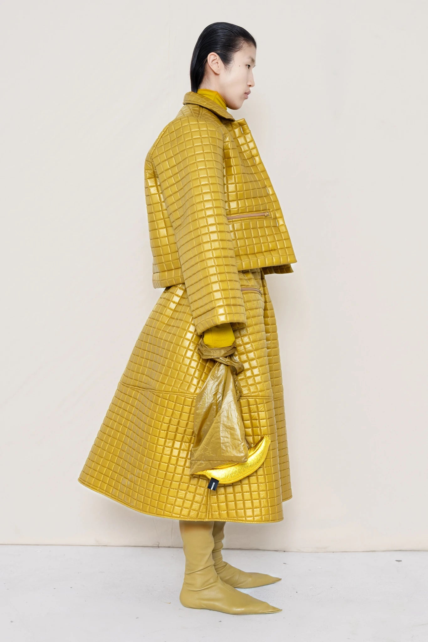 PERSON-WEARING-GOLD-YELLOW-SLEEVE-MOCK-NECK-TOP-WITH-MATCHING-QUILTEDJACKET-WITH-COLLAR-AND-A-LINE-SKIRT-ACCESSORIZED-WITH-MB-CHARM-NECKLACE-AND-MUD-COLORED-POINTY-FLAT-BOOTS-HOLDING--MATCHING-BANANA-BAG-DETAIL-SIDE-VIEW