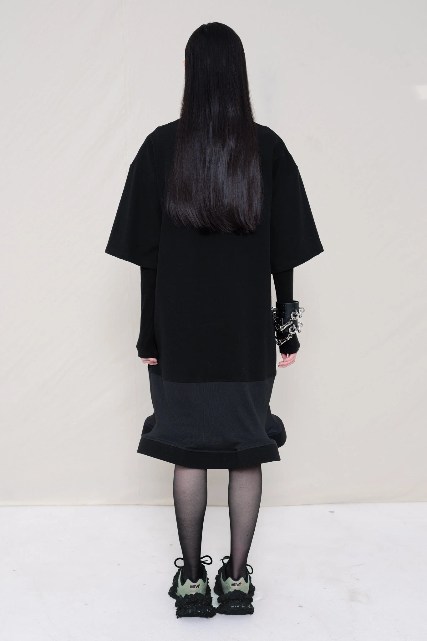 woman-wearing-A-black-T-SHIRT-STYLE-DRESS-WITH-A-WAVY-HEM-over-a-black-turtle-neck-paired-with-custom-shoes-and-black-leather-studded-bracelet-against-a-plain-background-full-body-side-view
