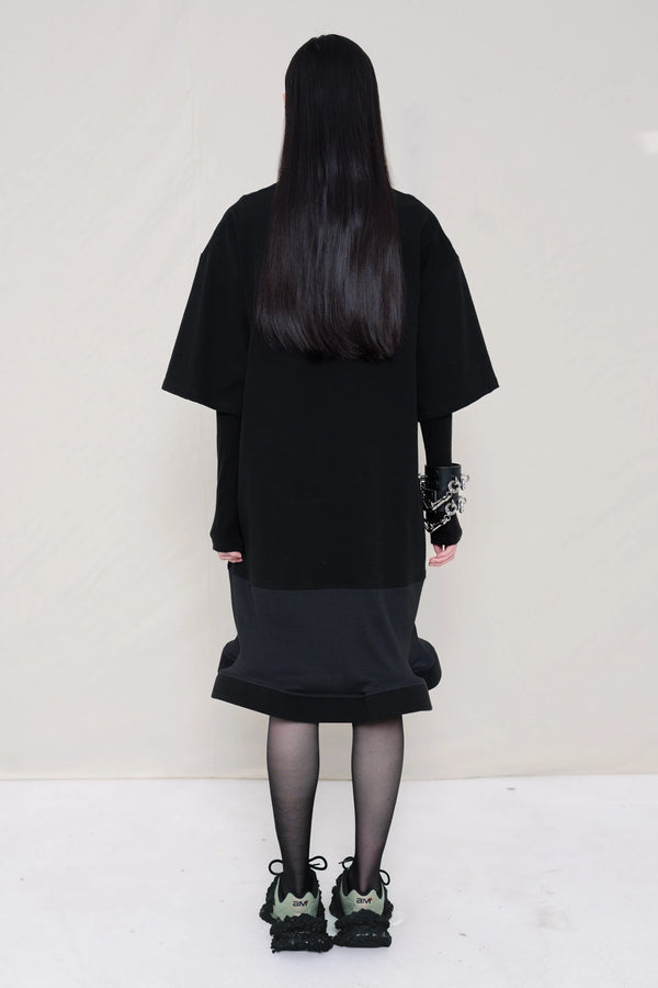 woman-wearing-A-black-T-SHIRT-STYLE-DRESS-WITH-A-WAVY-HEM-over-a-black-turtle-neck-paired-with-custom-shoes-and-black-leather-studded-bracelet-against-a-plain-background-full-body-side-view