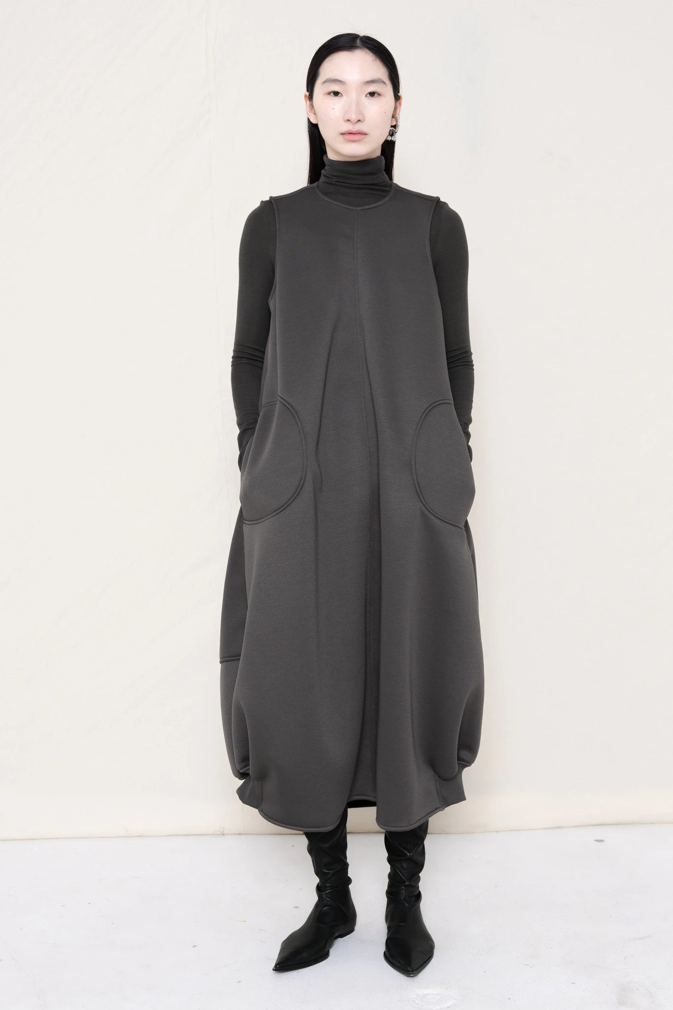 woman-wearing-a-grey-SLEEVELESS-O-LINE-SHAPED-DRESS-WITH-round-POCKETS-over-a-grey-turtle-neck-and-black-leather-pointed-boots-against a-white-background-full-body-front-view