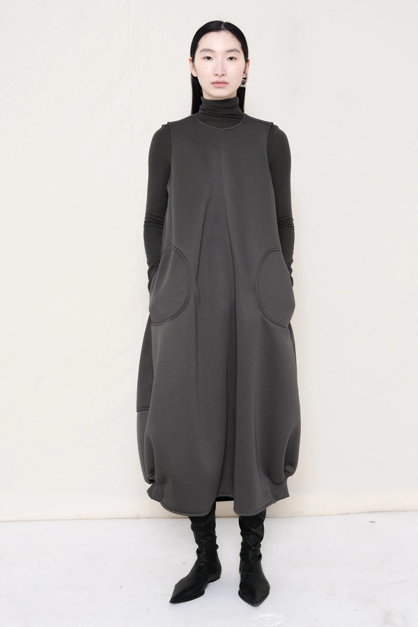 woman-wearing-a-grey-SLEEVELESS-O-LINE-SHAPED-DRESS-WITH-round-POCKETS-over-a-grey-turtle-neck-and-black-leather-pointed-boots-against a-white-background-full-body-front-view