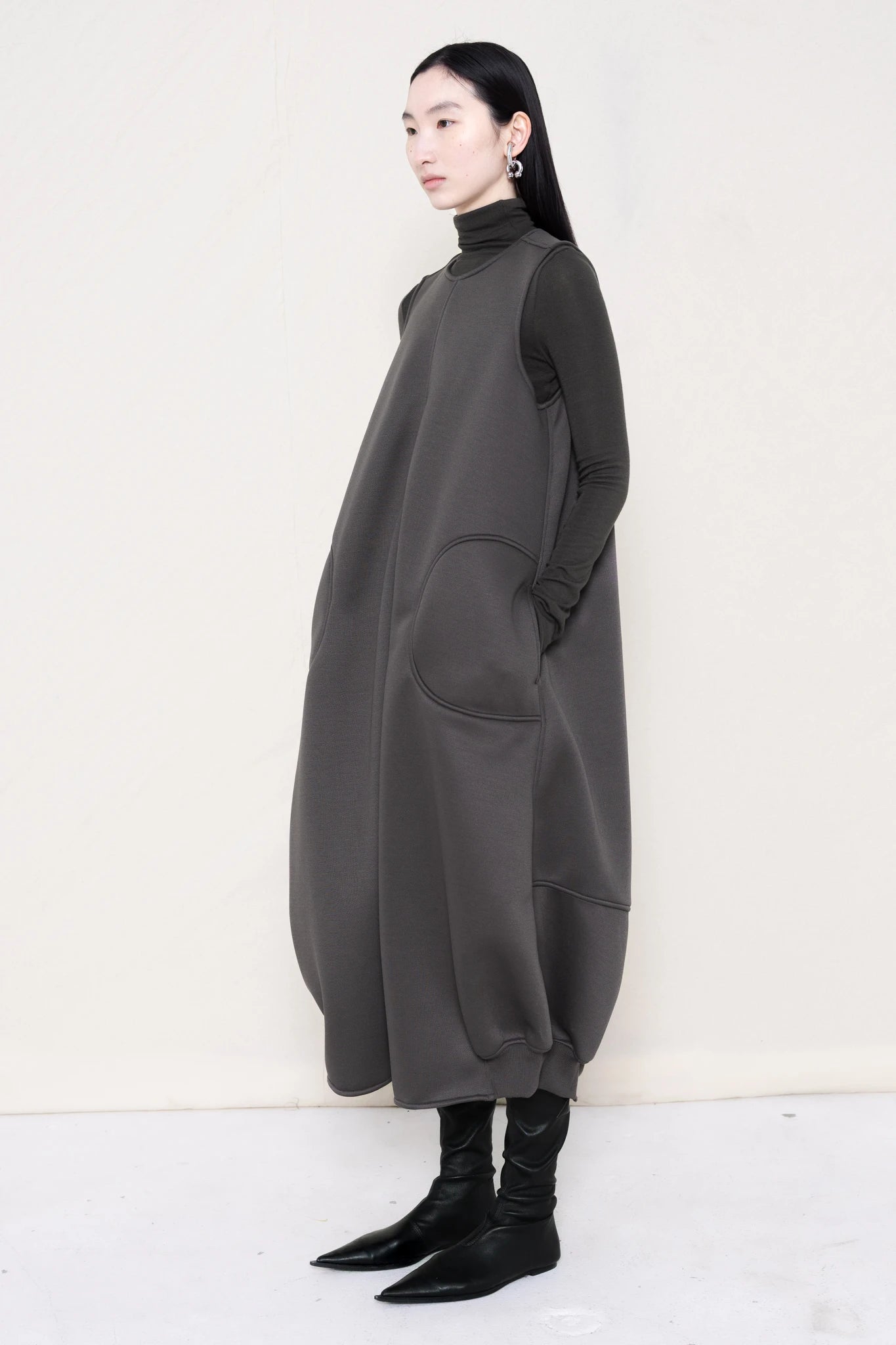woman-wearing-a-grey-SLEEVELESS-O-LINE-SHAPED-DRESS-WITH-round-POCKETS-over-a-grey-turtle-neck-and-black-leather-pointed-boots-against a-white-background-full-body-three-quarters-view