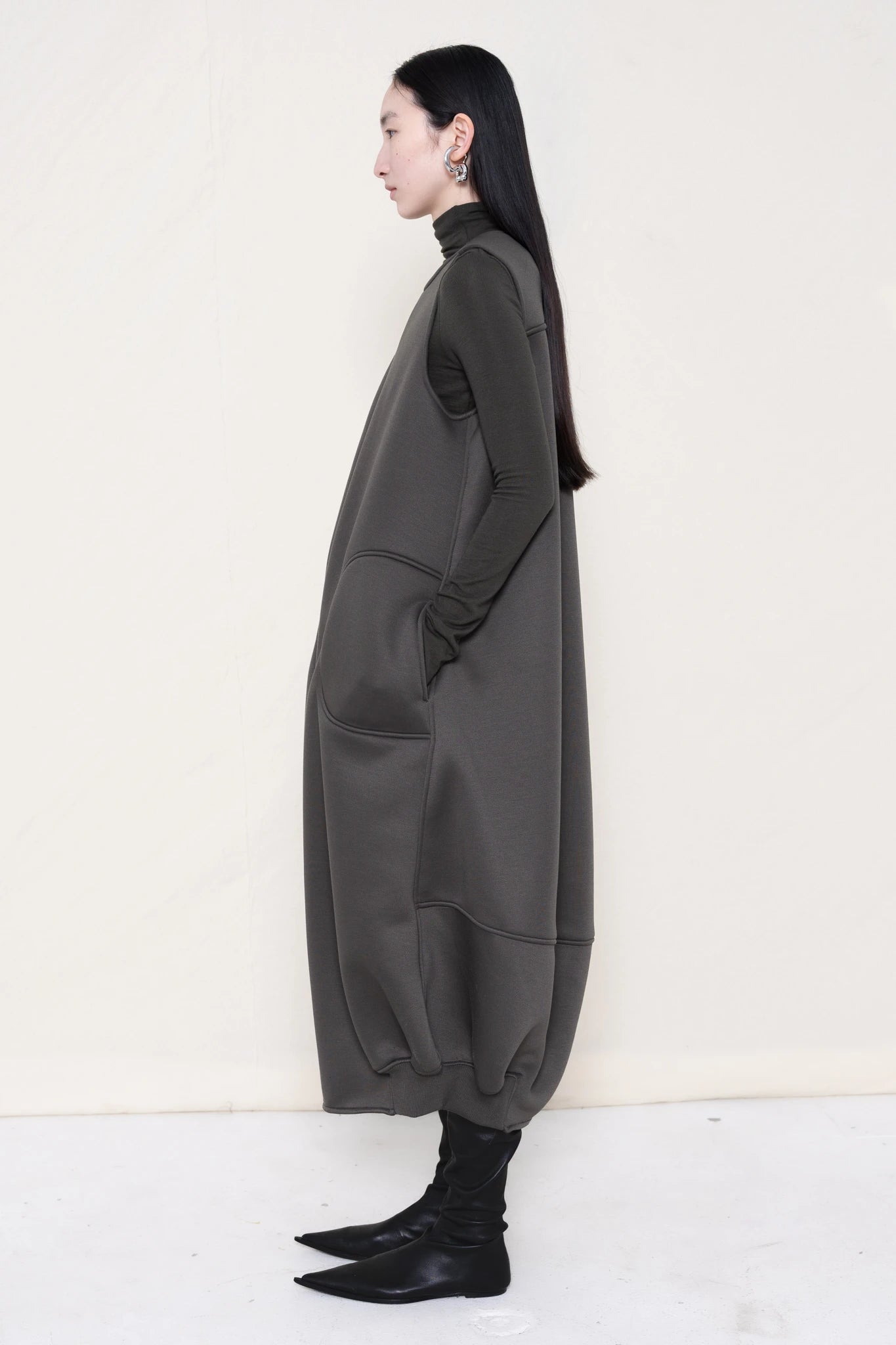 woman-wearing-a-grey-SLEEVELESS-O-LINE-SHAPED-DRESS-WITH-round-POCKETS-over-a-grey-turtle-neck-and-black-leather-pointed-boots-against a-white-background-full-body-side-view