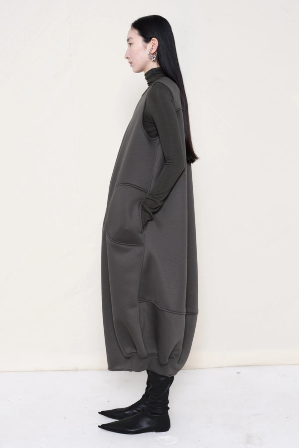woman-wearing-a-grey-SLEEVELESS-O-LINE-SHAPED-DRESS-WITH-round-POCKETS-over-a-grey-turtle-neck-and-black-leather-pointed-boots-against a-white-background-full-body-side-view