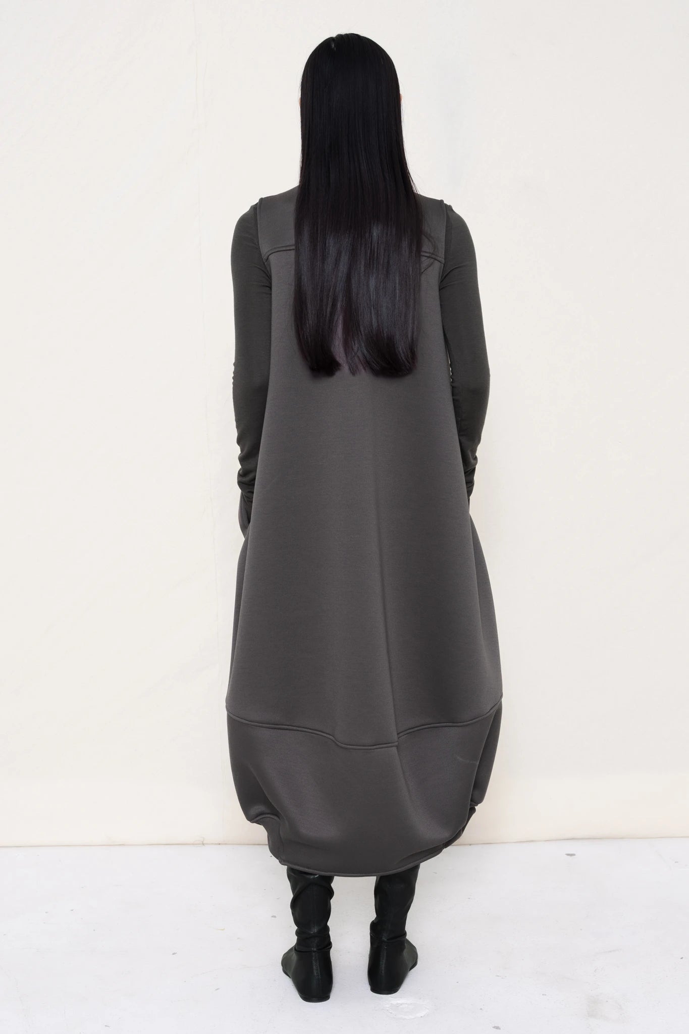 woman-wearing-a-grey-SLEEVELESS-O-LINE-SHAPED-DRESS-WITH-round-POCKETS-over-a-grey-turtle-neck-and-black-leather-pointed-boots-against a-white-background-full-body-back-view
