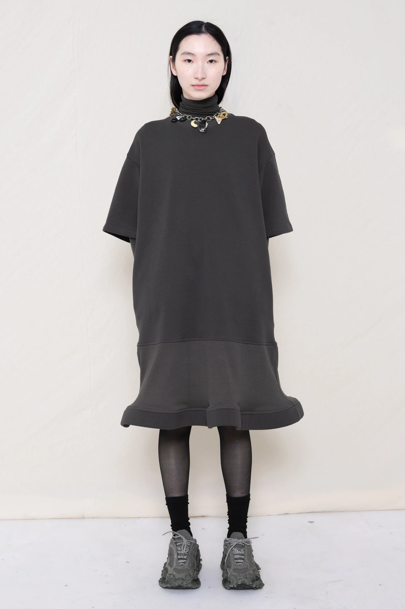 woman-wearing-A-grey-T-SHIRT-STYLE-DRESS-WITH-A-WAVY-HEM-over-a-grey-turtle-neck-paired-with-custom-grey-shoes-and-chain-chocker-against-a-plain-background-full-body-front-view