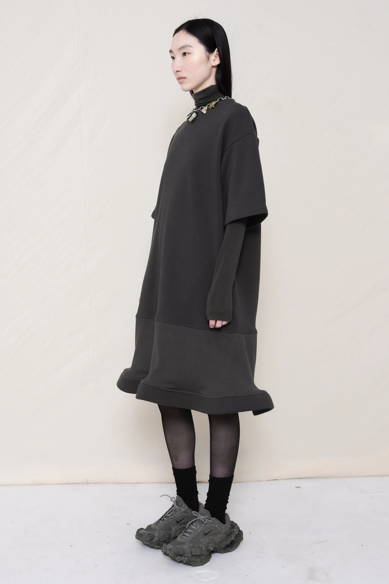 woman-wearing-A-grey-T-SHIRT-STYLE-DRESS-WITH-A-WAVY-HEM-over-a-grey-turtle-neck-paired-with-custom-grey-shoes-and-chain-chocker-against-a-plain-background-full-body-three-quarters-view