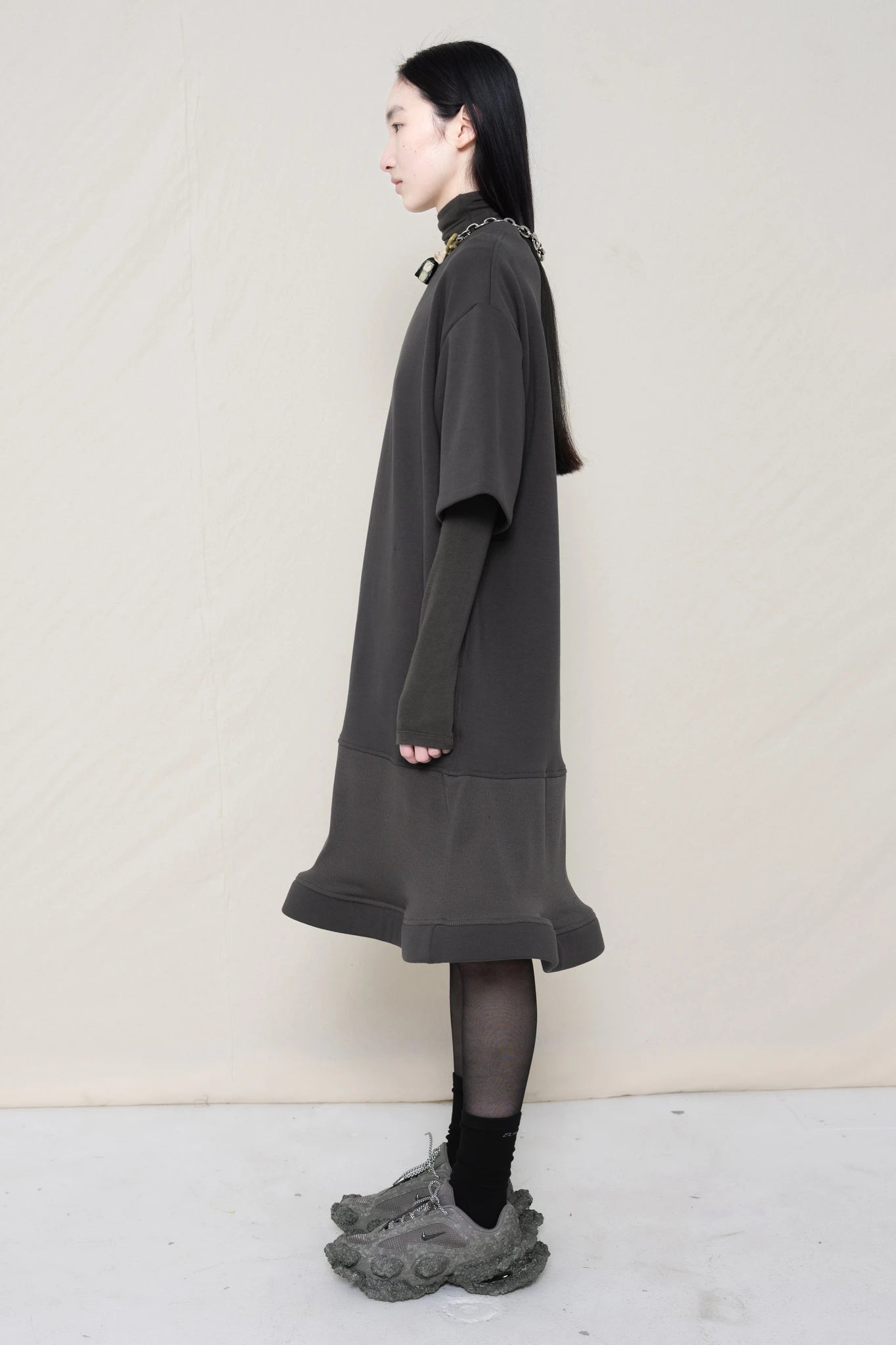 woman-wearing-A-grey-T-SHIRT-STYLE-DRESS-WITH-A-WAVY-HEM-over-a-grey-turtle-neck-paired-with-custom-grey-shoes-and-chain-chocker-against-a-plain-background-full-body-side-view