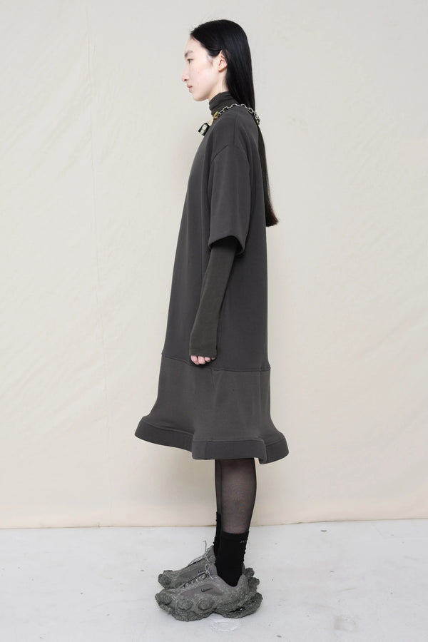 woman-wearing-A-grey-T-SHIRT-STYLE-DRESS-WITH-A-WAVY-HEM-over-a-grey-turtle-neck-paired-with-custom-grey-shoes-and-chain-chocker-against-a-plain-background-full-body-side-view