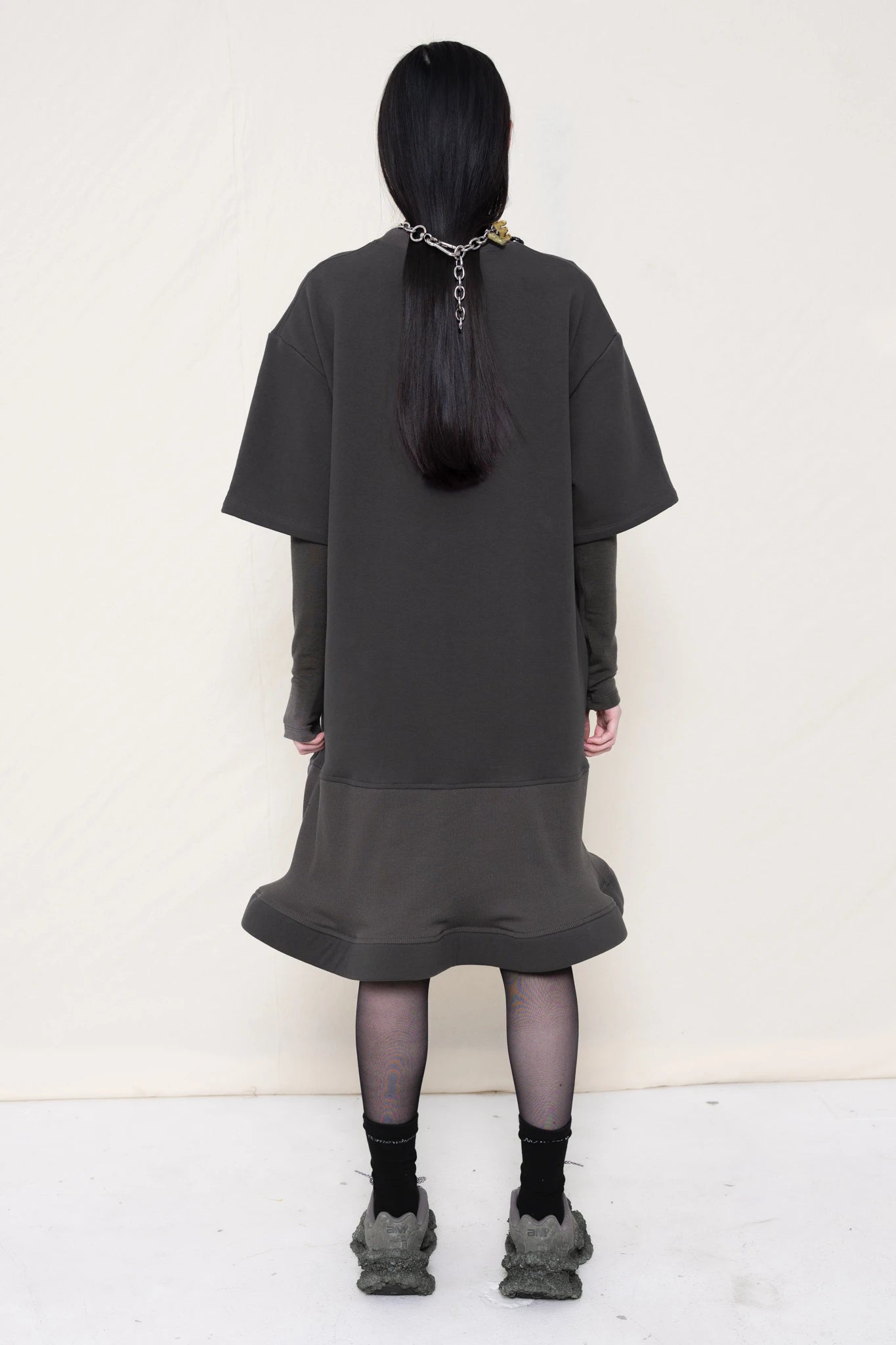 woman-wearing-A-grey-T-SHIRT-STYLE-DRESS-WITH-A-WAVY-HEM-over-a-grey-turtle-neck-paired-with-custom-grey-shoes-and-chain-chocker-against-a-plain-background-full-body-back-view