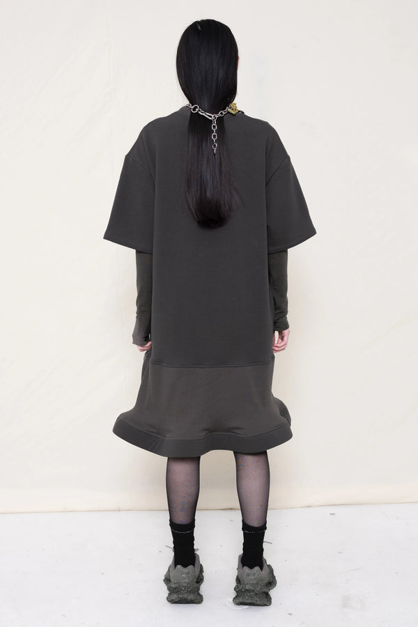 woman-wearing-A-grey-T-SHIRT-STYLE-DRESS-WITH-A-WAVY-HEM-over-a-grey-turtle-neck-paired-with-custom-grey-shoes-and-chain-chocker-against-a-plain-background-full-body-back-view