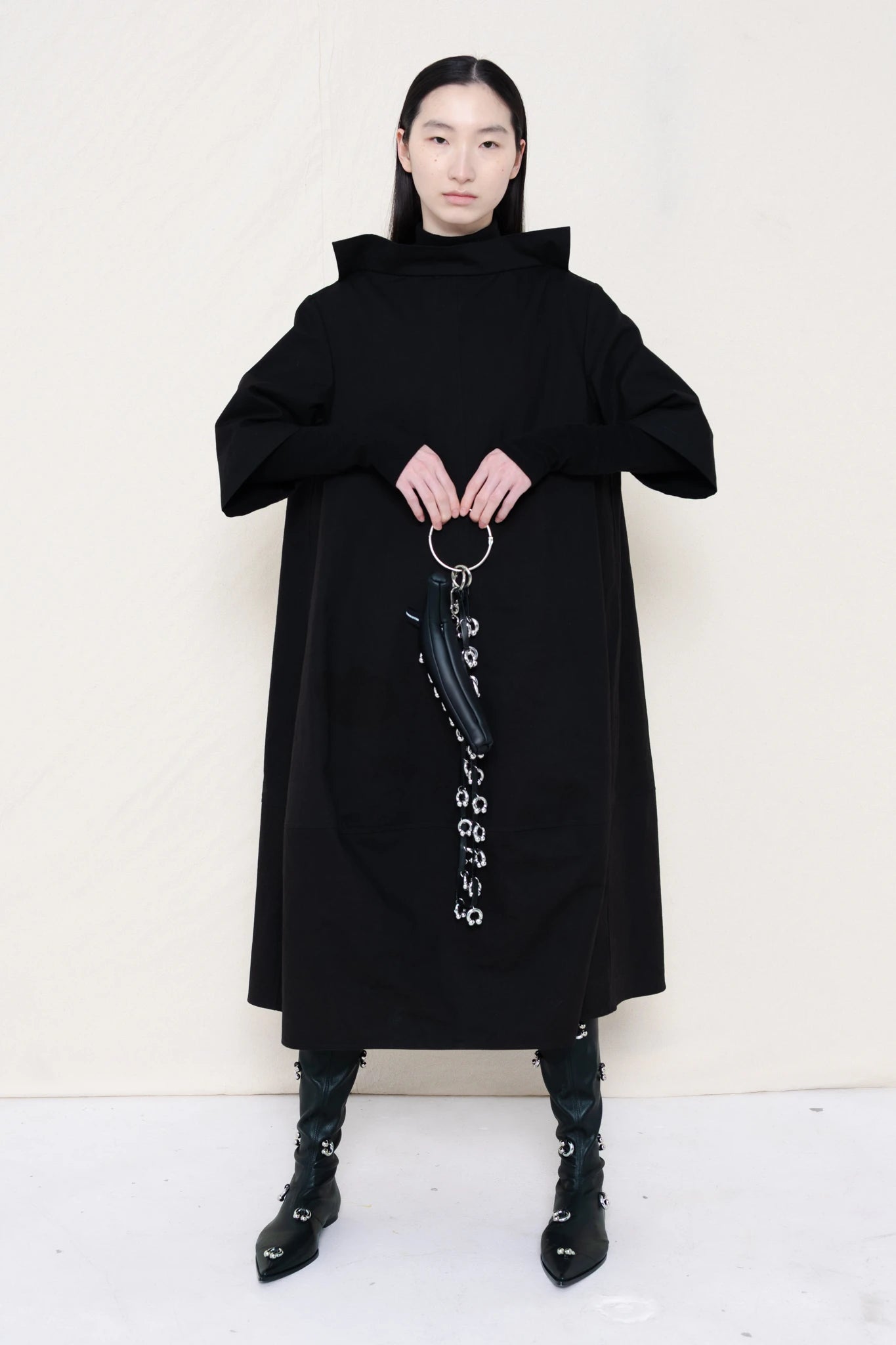 woman-holding-a-black-leather-studded-banana-wearing-a-calf-length-black-a-line-boatneck-dress-with-mid-length-sleeves-worn-over-a-black-turtle-neck-worn-with-black-pointed-studded-leather-boots-against-a-plain-background-full-body-front-view