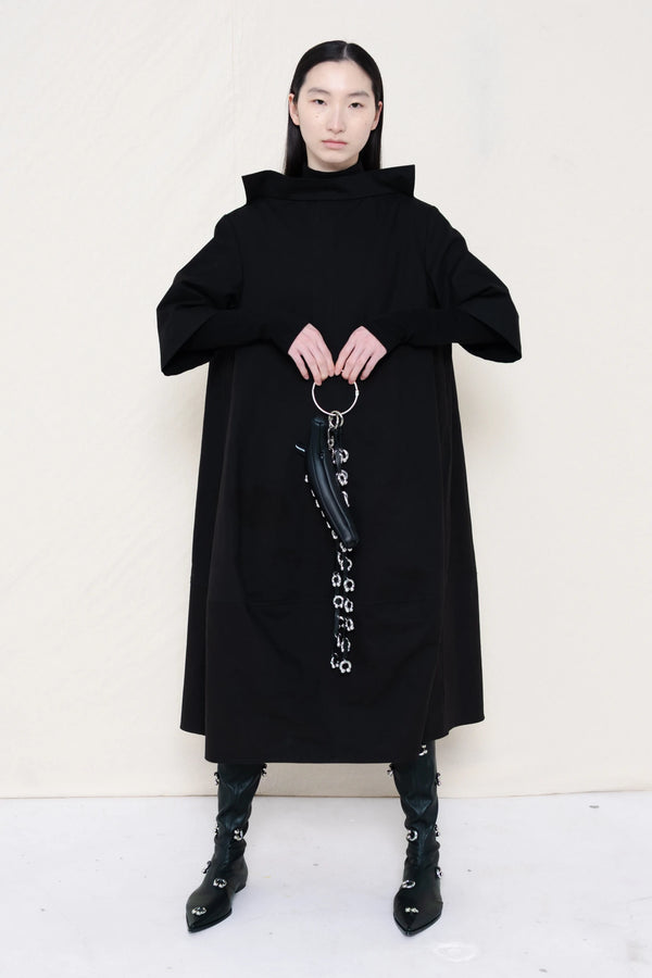 woman-holding-a-black-leather-studded-banana-wearing-a-calf-length-black-a-line-boatneck-dress-with-mid-length-sleeves-worn-over-a-black-turtle-neck-worn-with-black-pointed-studded-leather-boots-against-a-plain-background-full-body-front-view