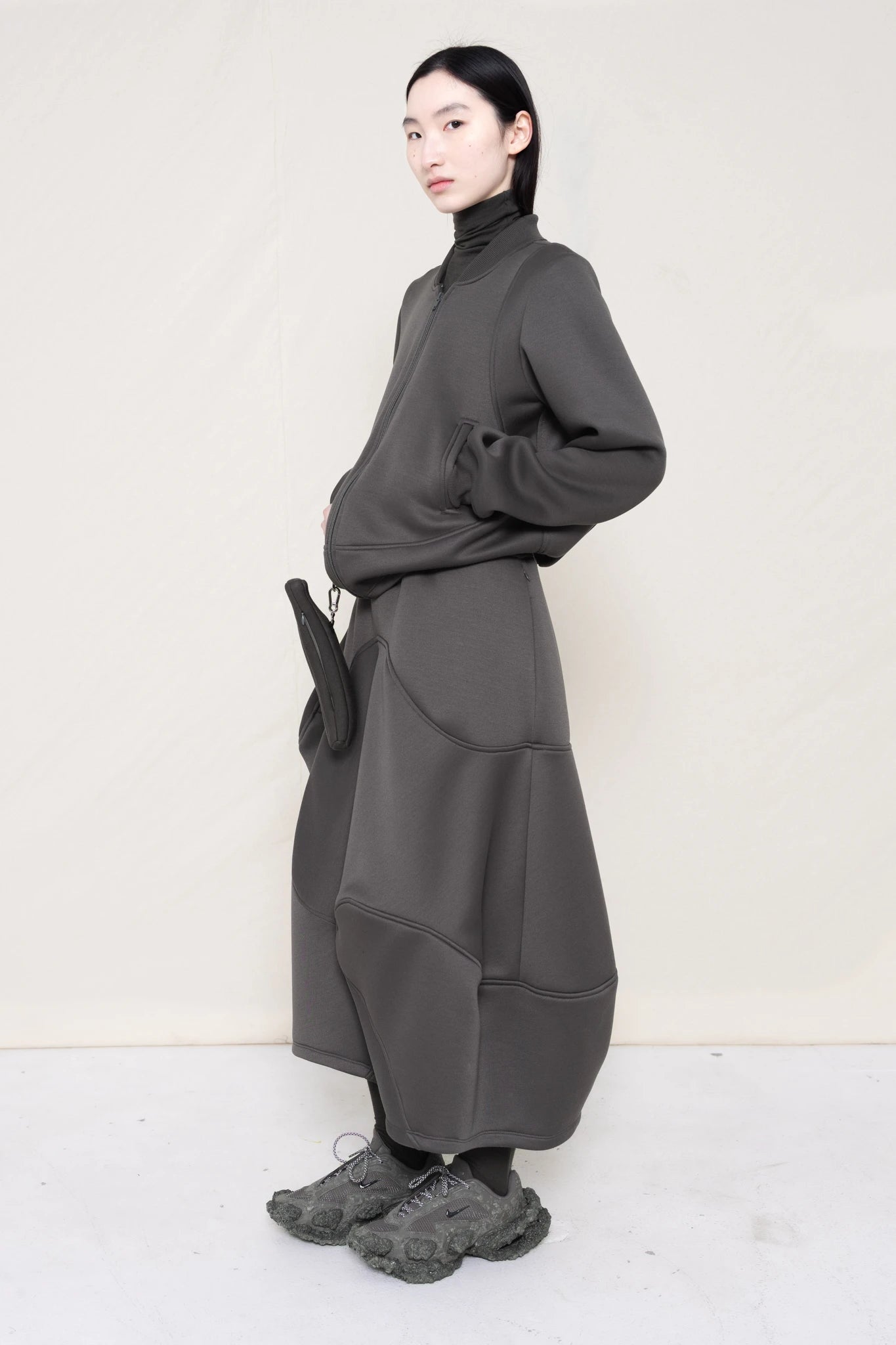 PERSON-WEARING-STONE-GREY-WAVY-ZIP-UP-WITH-MATCHING-WAVY-SEAM-SKIRT-ACCESSORIZED-WITH-CUSTOM-SNEAKERS-AND-GREY-BANANA-TOTE-BAG-THREE-QUARTER-VIEW