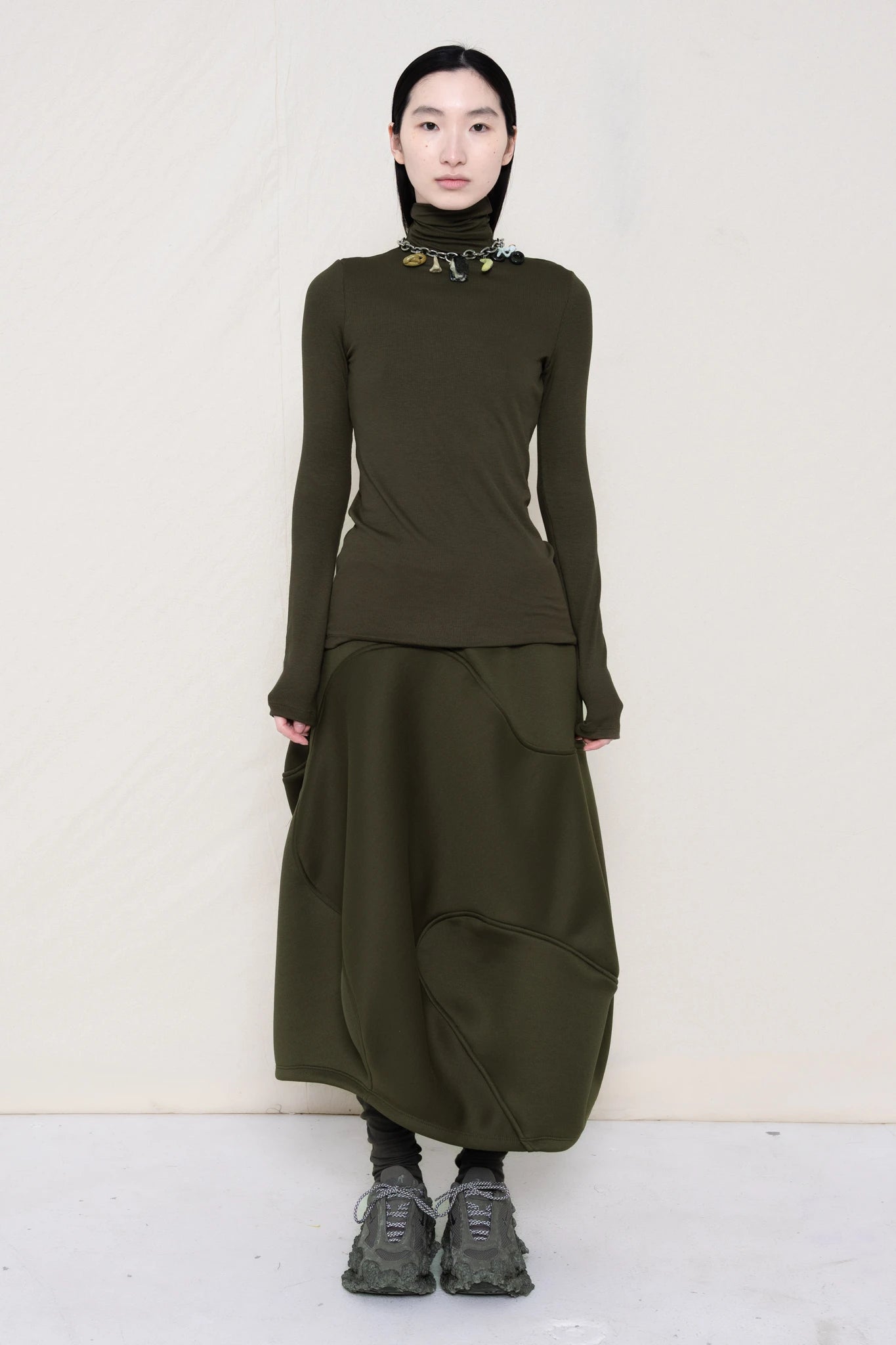 woman-wearing-a-cold-moss-green-soft-fluid-rib-long-sleeve-turtleneck-with-an-mb-charm-necklace-over-a-moss-green-spongy-seamed-skirt-and-custom-shoes-against-a-white-background-full-body-front-view