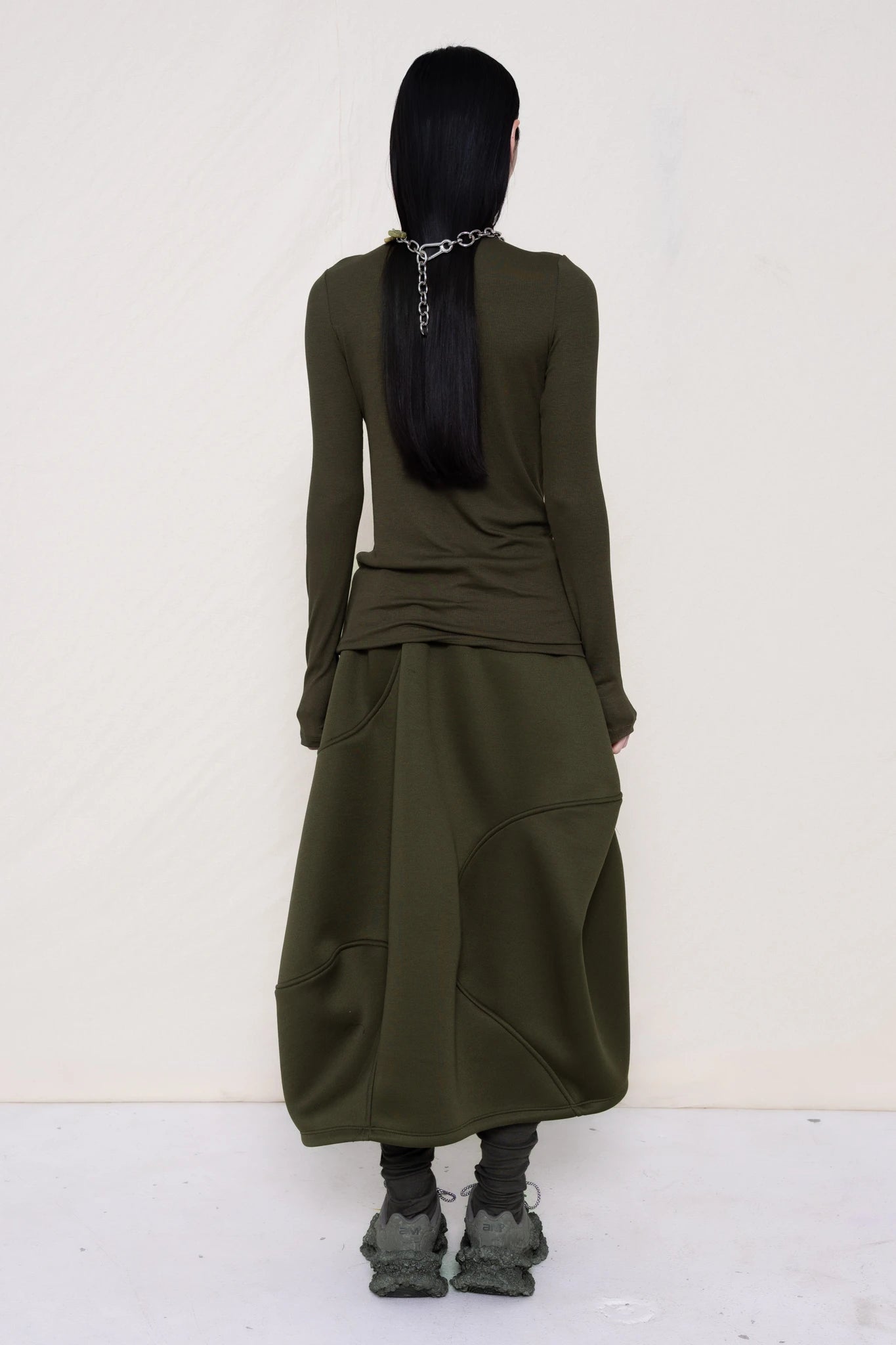 woman-wearing-a-cold-moss-green-soft-fluid-rib-long-sleeve-turtleneck-with-an-mb-charm-necklace-over-a-moss-green-spongy-seamed-skirt-and-custom-shoes-against-a-white-background-full-body-back-view