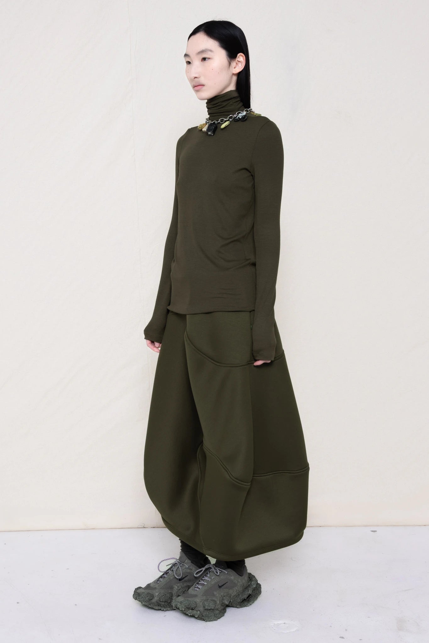 woman-wearing-a-cold-moss-green-soft-fluid-rib-long-sleeve-turtleneck-with-an-mb-charm-necklace-over-a-moss-green-spongy-seamed-skirt-and-custom-shoes-against-a-white-background-full-body-three-quarter-view
