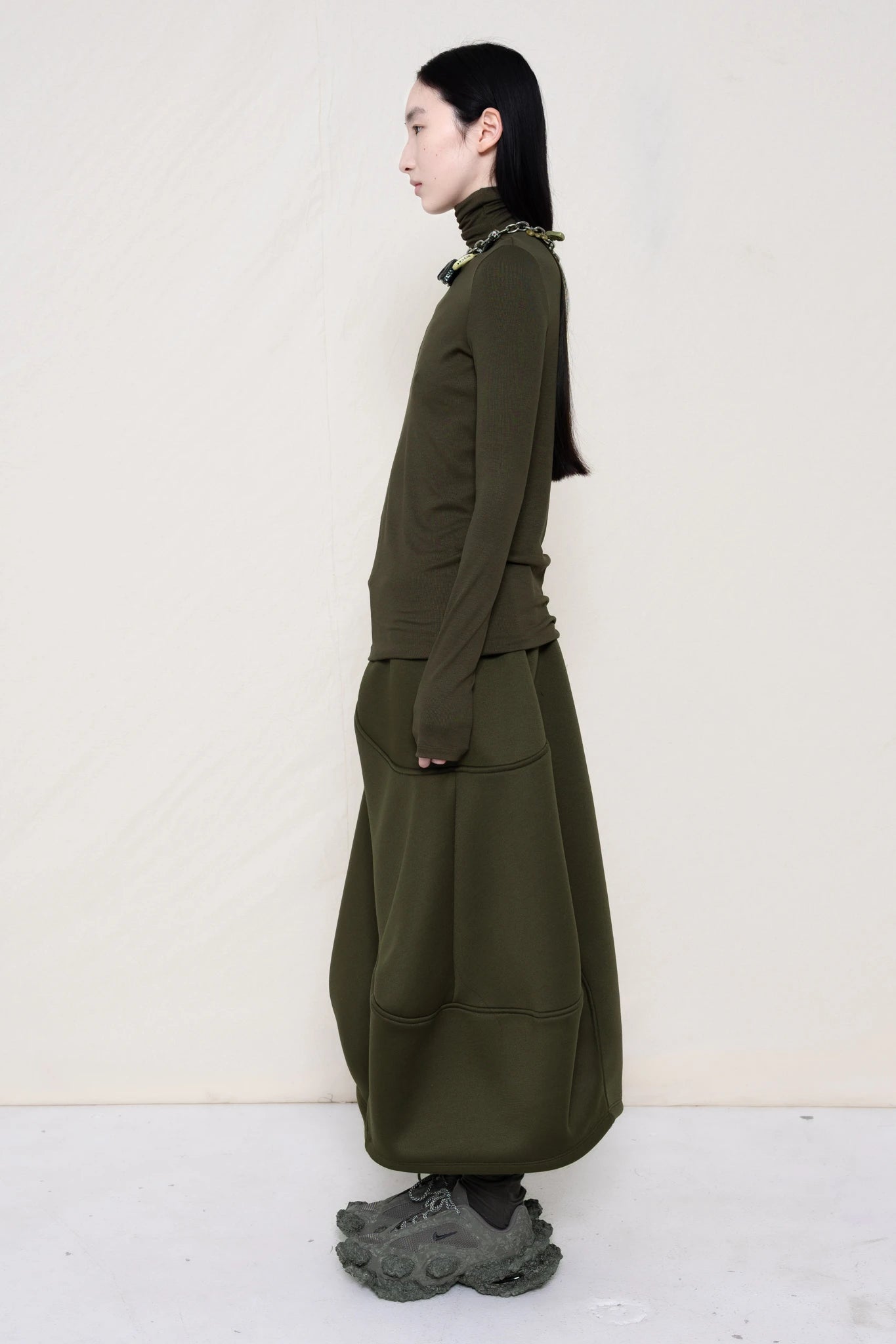 woman-wearing-a-cold-moss-green-soft-fluid-rib-long-sleeve-turtleneck-with-an-mb-charm-necklace-over-a-moss-green-spongy-seamed-skirt-and-custom-shoes-against-a-white-background-full-body-side-view