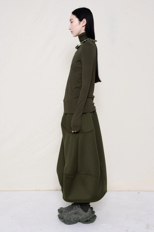 woman-wearing-a-cold-moss-green-soft-fluid-rib-long-sleeve-turtleneck-with-an-mb-charm-necklace-over-a-moss-green-spongy-seamed-skirt-and-custom-shoes-against-a-white-background-full-body-side-view