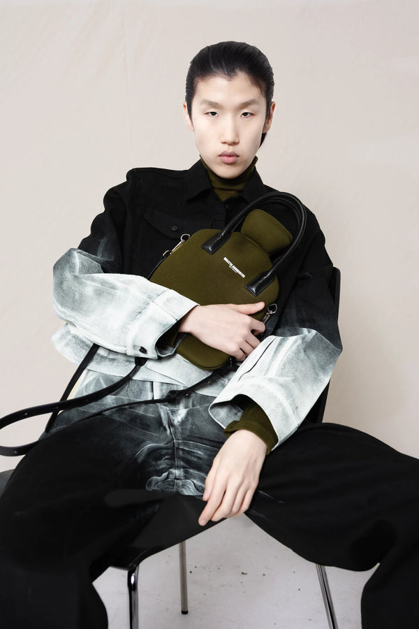 PERSON-WEARING-WHITE-OMBRE-PAINTED-JACKET-AND-PANTS-IN-BLACK-WITH-MOSS-UNDERSHIRT-SLOUCHING-IN-A-CHAIR-HOLDING-MOSS-COLORED-HOT-WATER-BOTTLE-SHAPED-BAG-WITH-LEATHER-HANDLES-FRONT-VIEW